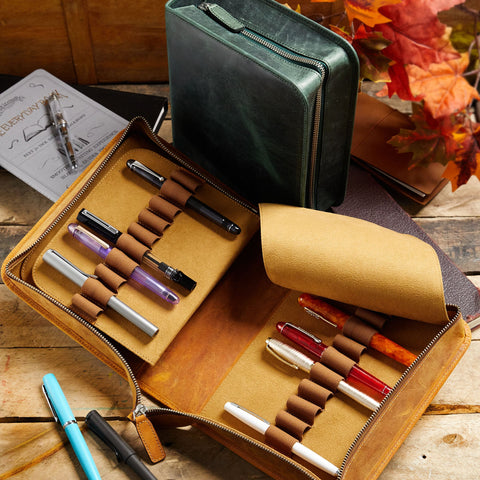 Galen Leather display with a green zippered case and a leather roll of colorful pens on a wooden surface.