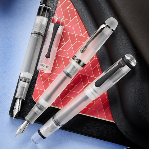 Eyedropper fountain pens with translucent bodies and metal nibs spread on a red notebook and blue backdrop.