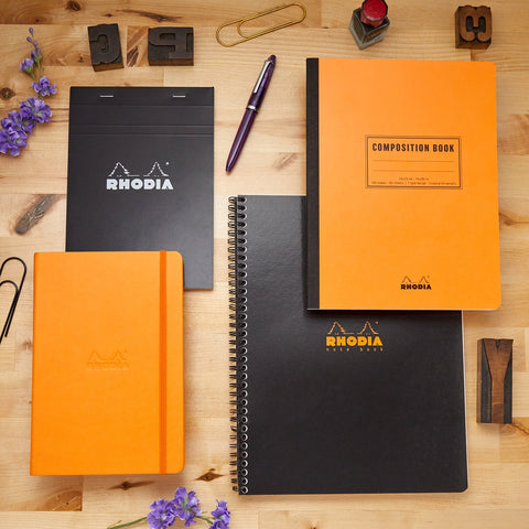 Rhodia notebooks in orange and black covers on a wooden desk with a purple pen.
