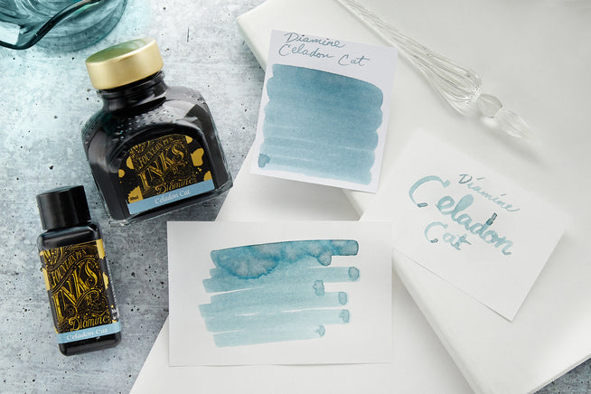 Diamine Celedon Cat Ink bottels, swabs and a glass dip pen on white paper and light blue background
