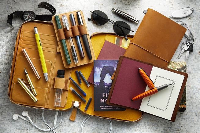 Assorted fountain pens on brown notebook with other items in the background