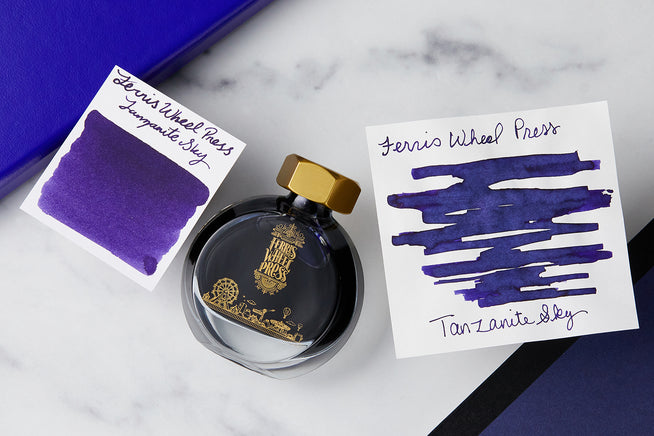 Ferris Wheel Press Tanzanite Sky fountain pen ink bottle, with 2 white swap cards with ink swas on them, on a marble background with a purple journal
