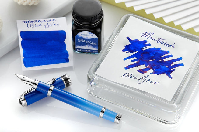 Monteverde Innova Blue sky fountain pen uncapped with bottle of blue skies ink, and 2 swabs of the ink on a white desktop bacground with some elevations to create visual interest. 