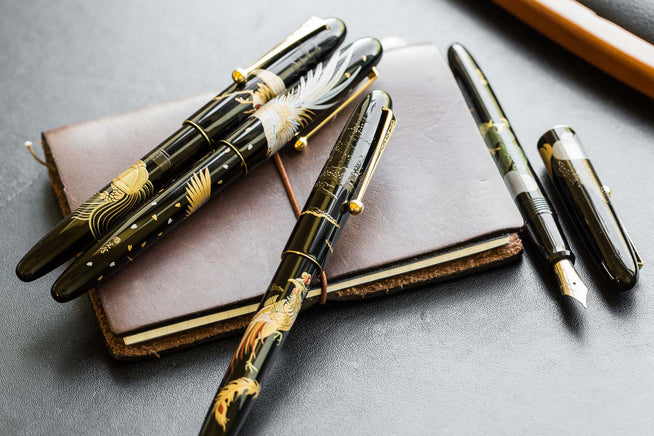 Group of assorted Namiki Nippon Art Maki-e fountain pens on dark gray backgroun, sitting on top of a brown leather journal
