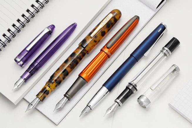 A group of five fountain pens diagonally lined up on open notebooks