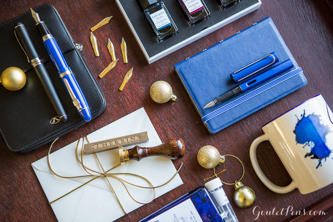 Assorted fountain pens along with nibs, cups, notebooks, and stationary sets on a wooden background with gold ornametns