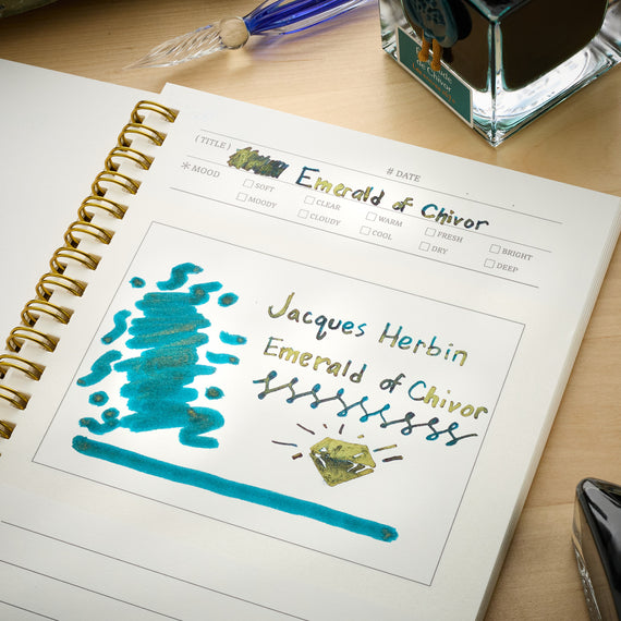 An open spiral notebook on a wooden desk showing a swatch page for the ink color 'Emerald of Chivor' by Jacques Herbin. The page features a blue-green ink swatch and handwritten notes on white paper, with an ink bottle and fountain pen visible in the background.